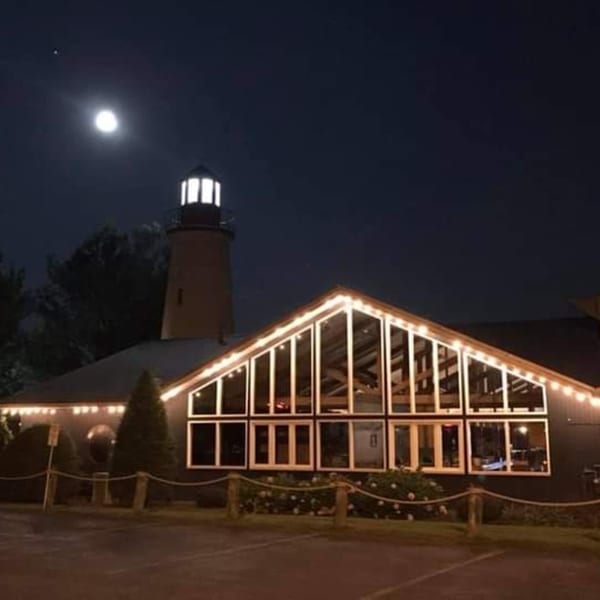 Lighthouse Bistro at night, with a full moon, illuminated by string lights and a glowing lighthouse.